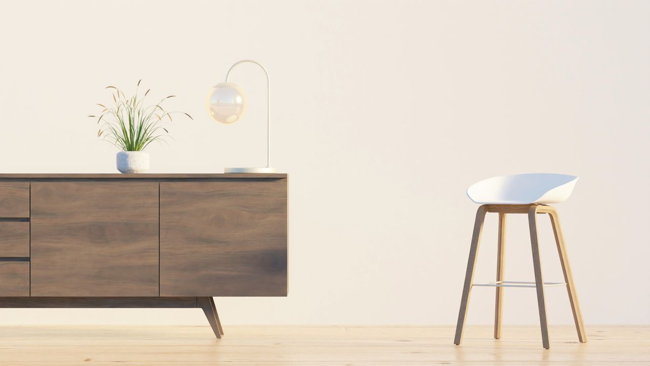 Modern minimalist interior featuring a wooden sideboard, chair, lamp, and plant against a white wall.