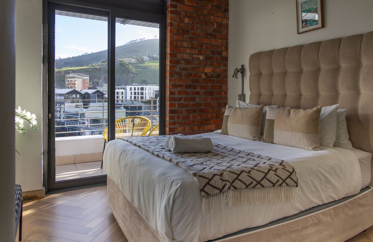 Modern bedroom with plush bed and balcony offering scenic views of hills and urban landscape.